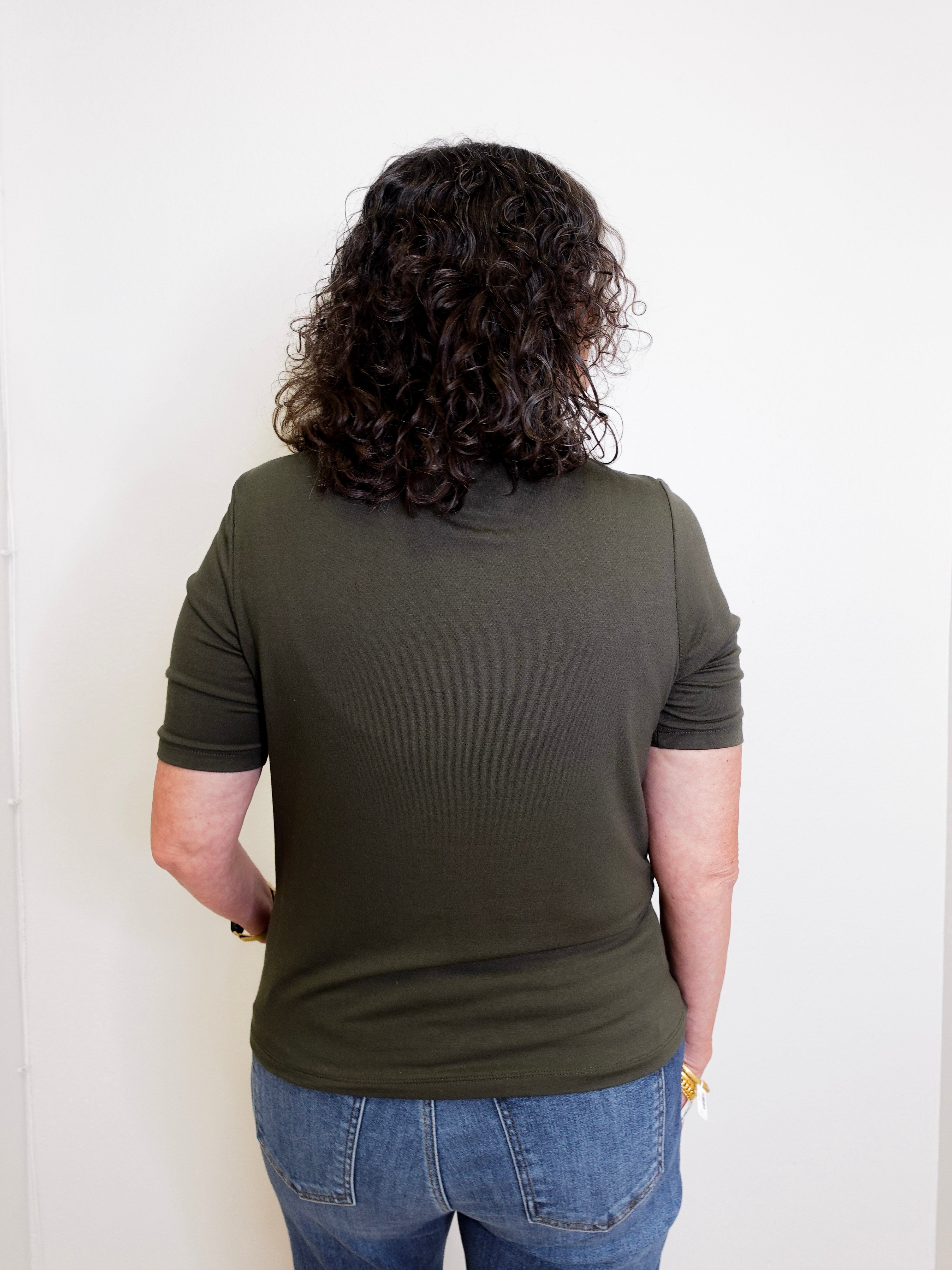 Person wearing an olive green t-shirt and blue jeans standing against a white wall.