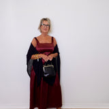 Woman in a burgundy dress with a black shawl holding a handbag against a white wall.
