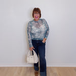 Woman wearing a patterned long-sleeve top, jeans, and brown shoes, holding a white handbag against a plain background.