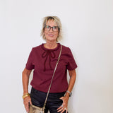 Woman wearing a burgundy top with a bow, black pants, and holding a gold handbag against a white background.