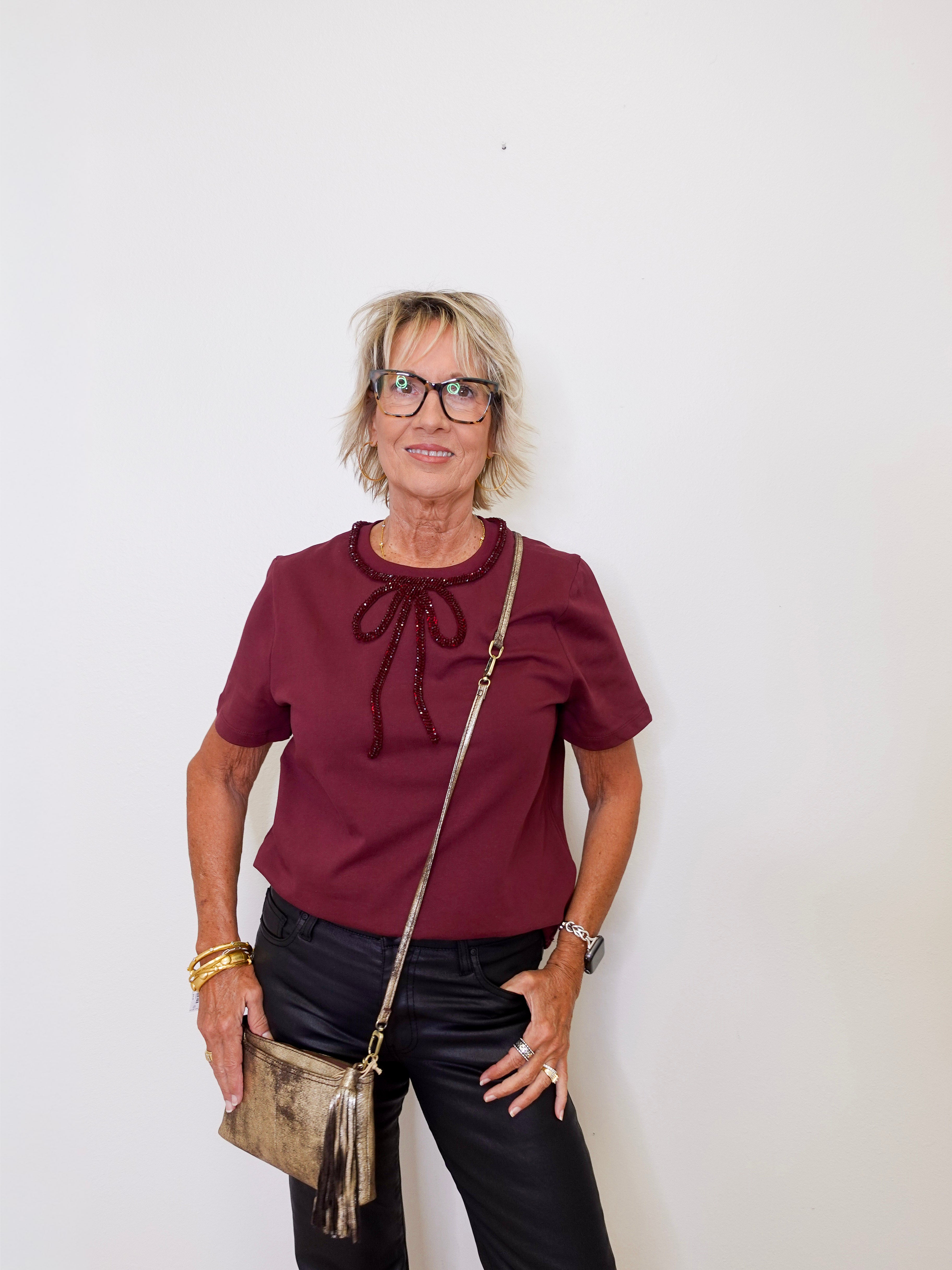 Woman wearing a burgundy top with a bow, black pants, and holding a gold handbag against a white background.