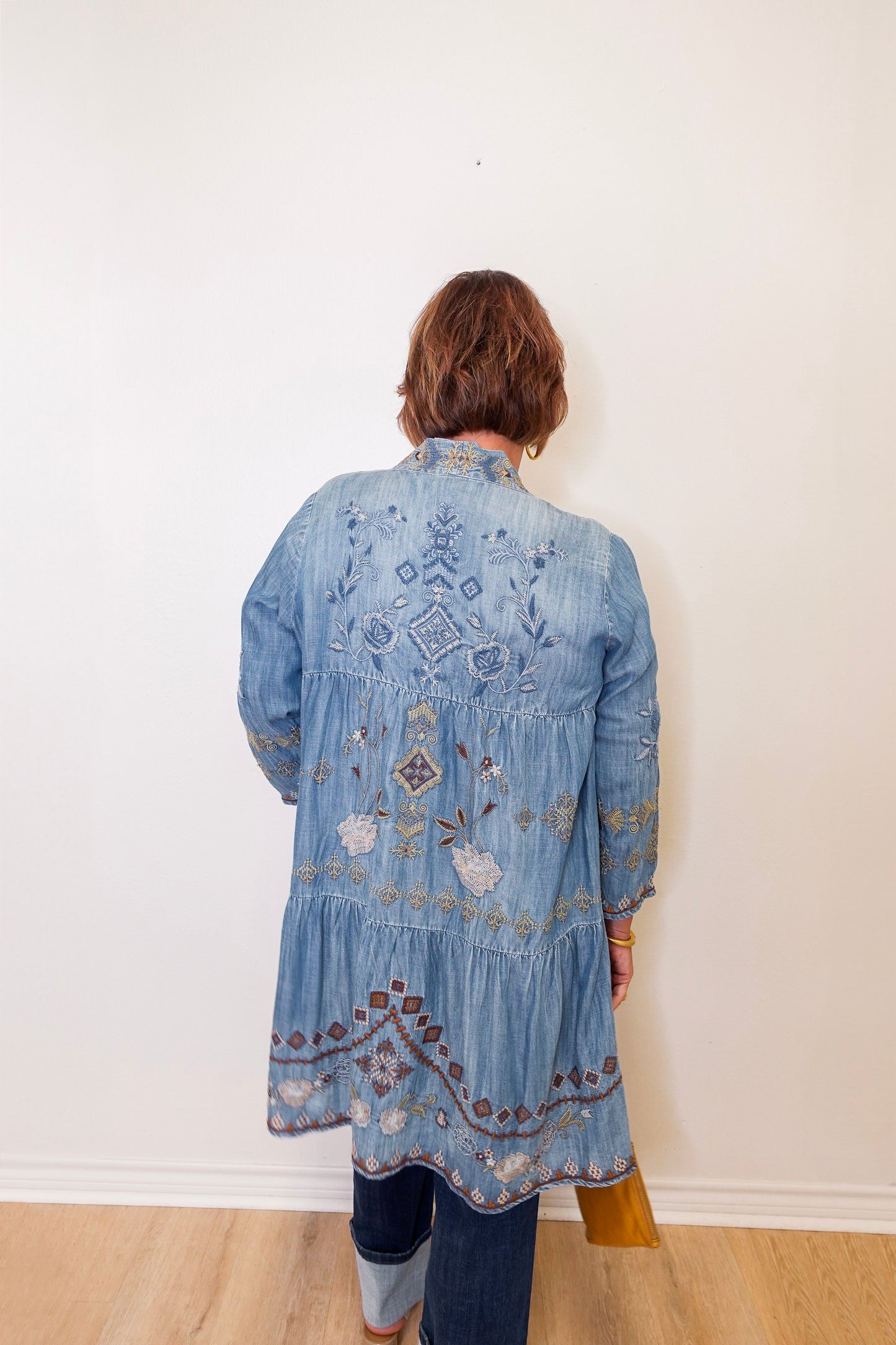 Person wearing a blue embroidered denim jacket against a white wall.