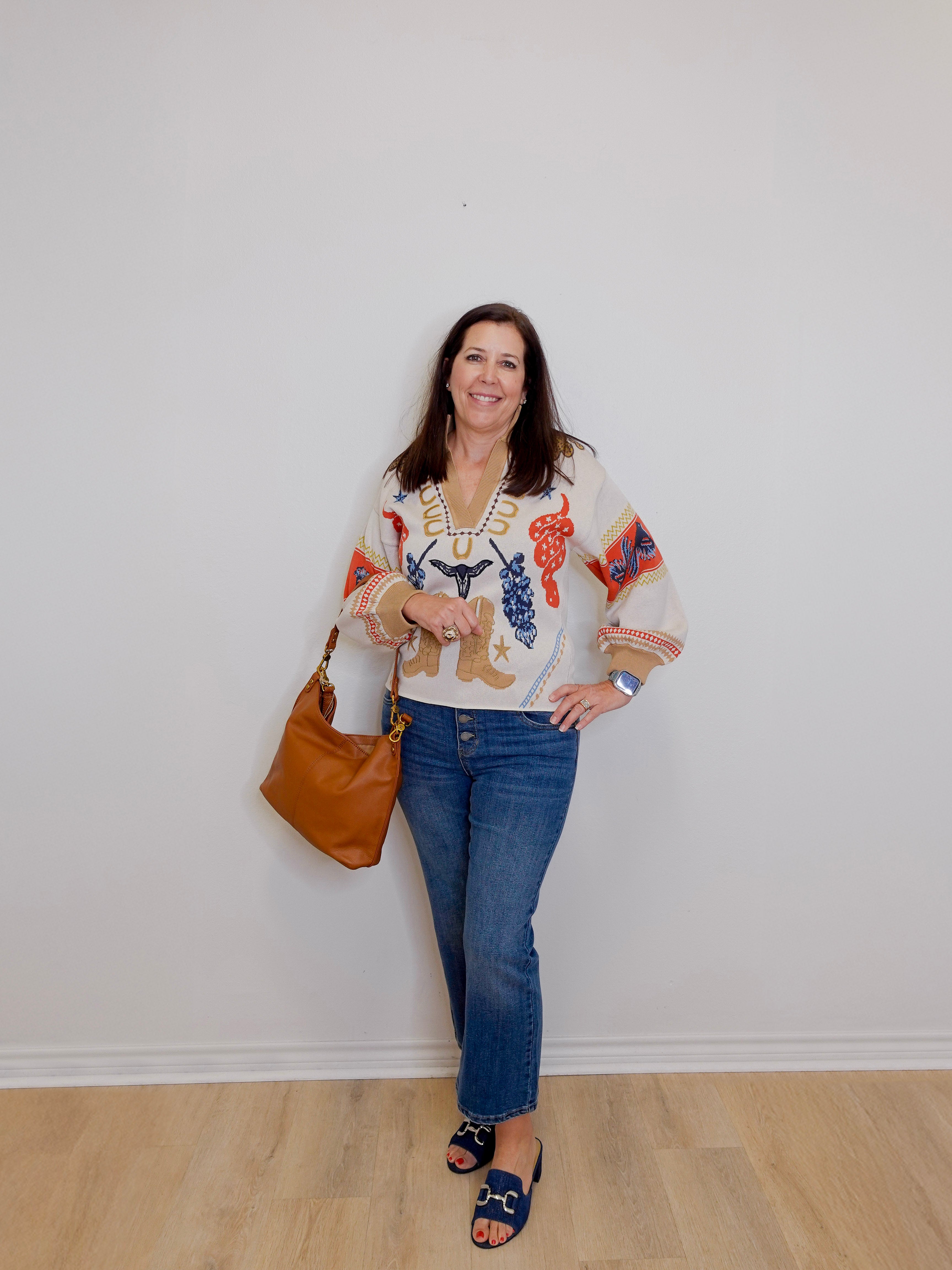 Woman wearing a patterned blouse and jeans, holding a brown handbag against a white wall.