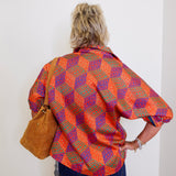 Person wearing a colorful patterned shirt and blue jeans, holding a brown leather bag against a white wall.