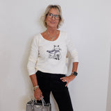 Woman wearing a white sweater with a cat design, holding a silver handbag against a white wall.