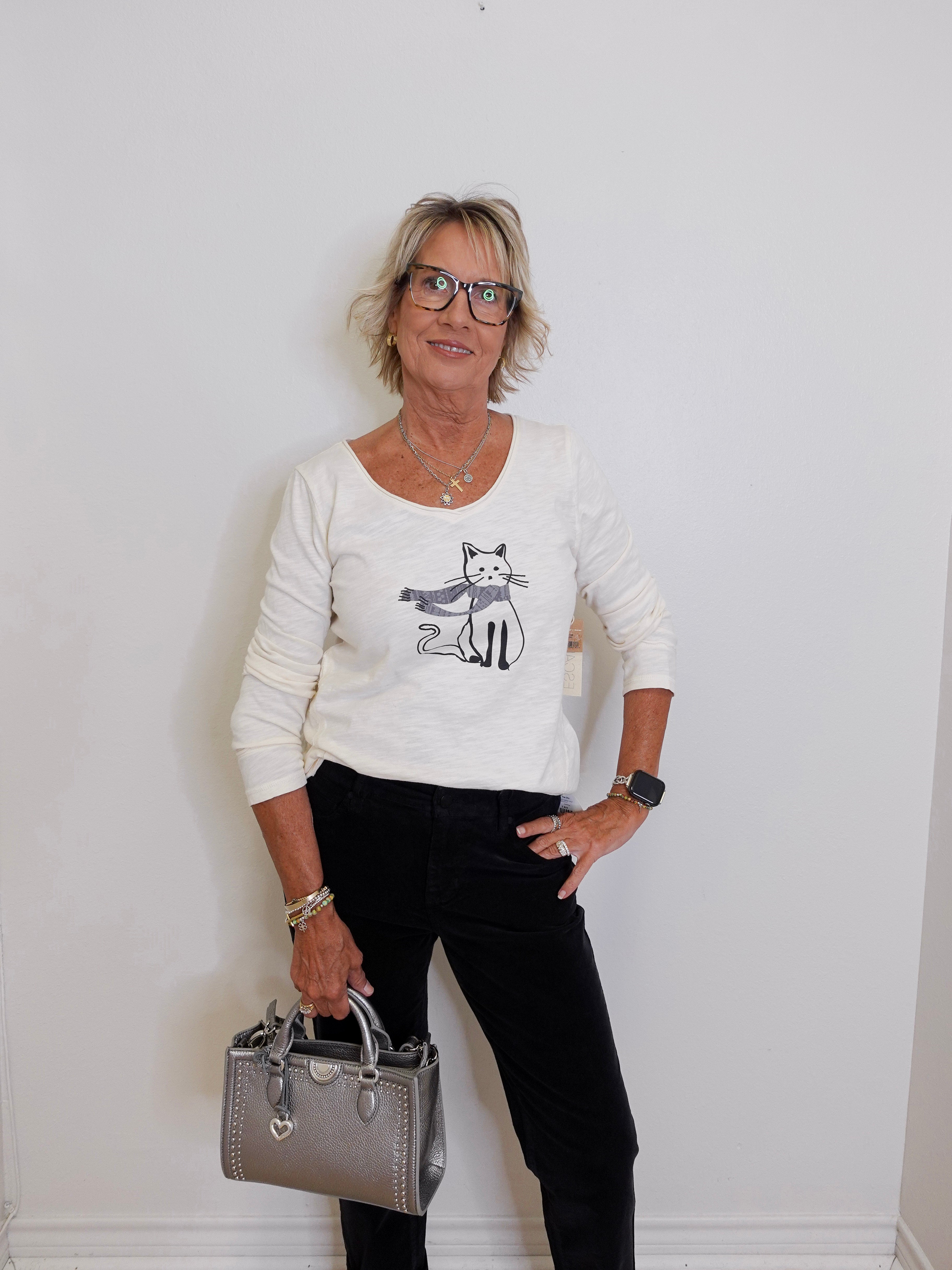 Woman wearing a white sweater with a cat design, holding a silver handbag against a white wall.