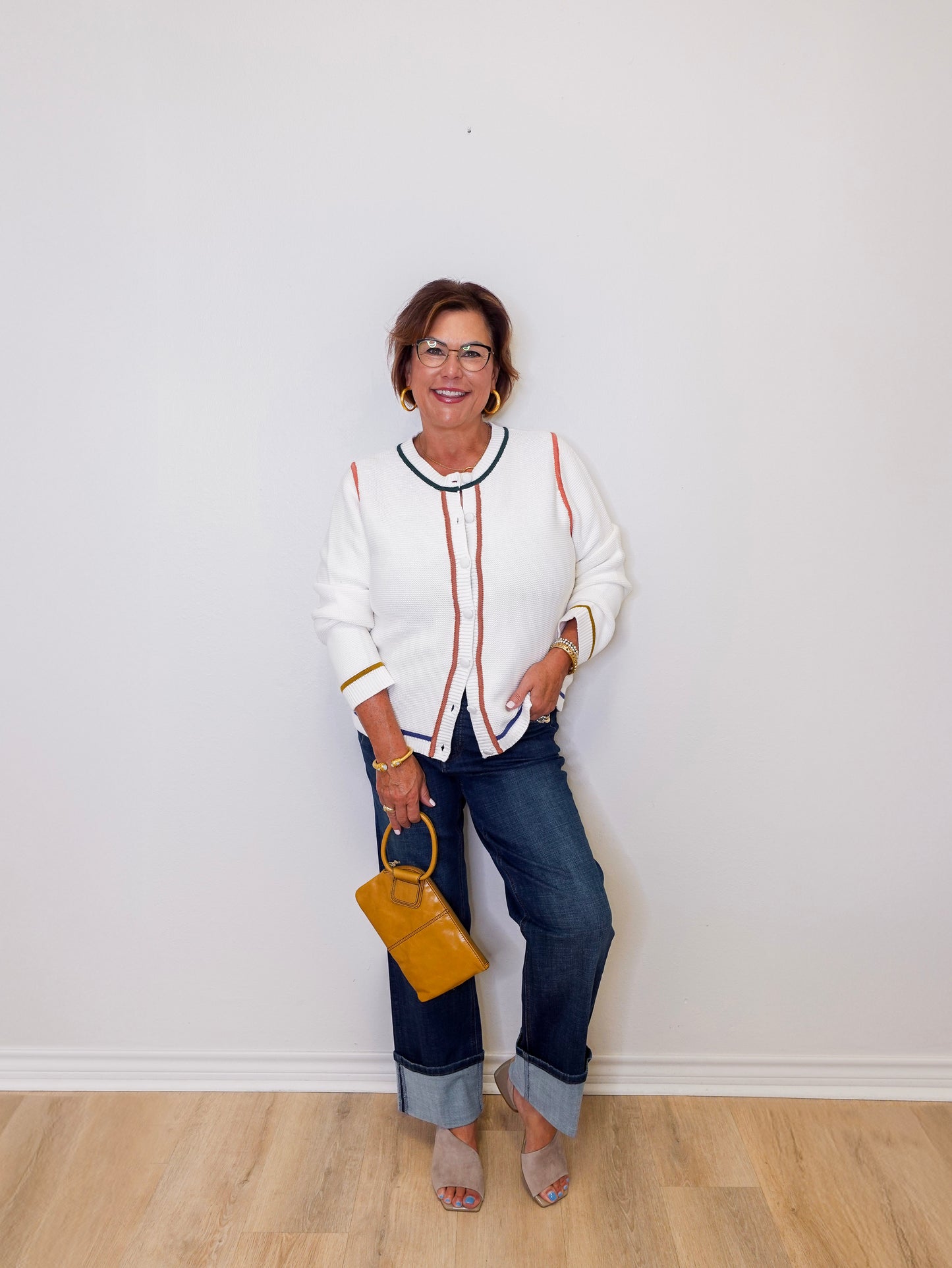 Woman wearing a white sweater with red accents and blue jeans, holding a yellow handbag against a white wall.