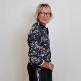 Woman wearing a black and white floral shirt and black pants against a plain white wall.