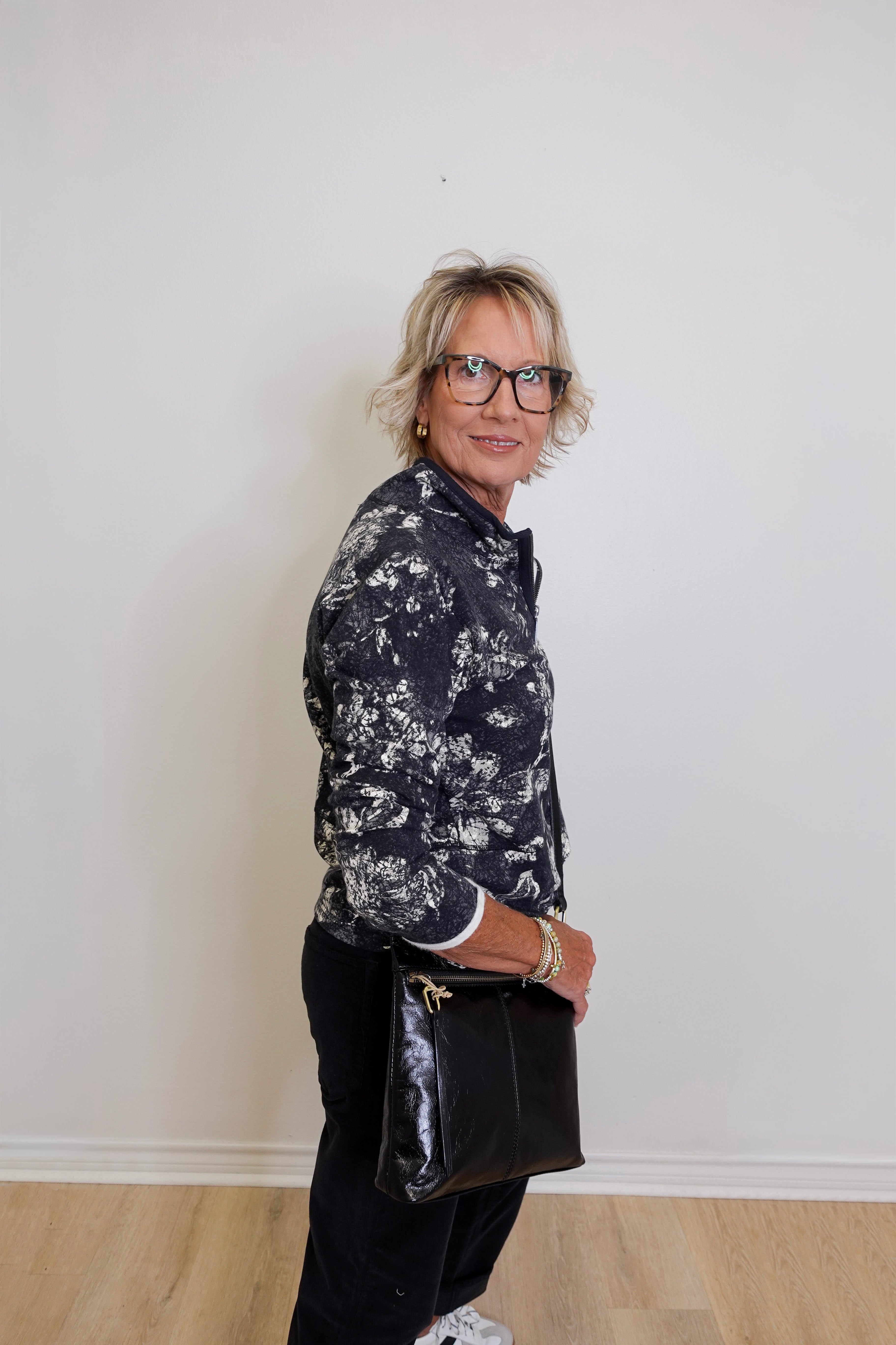 Woman wearing a black and white floral shirt and black pants against a plain white wall.