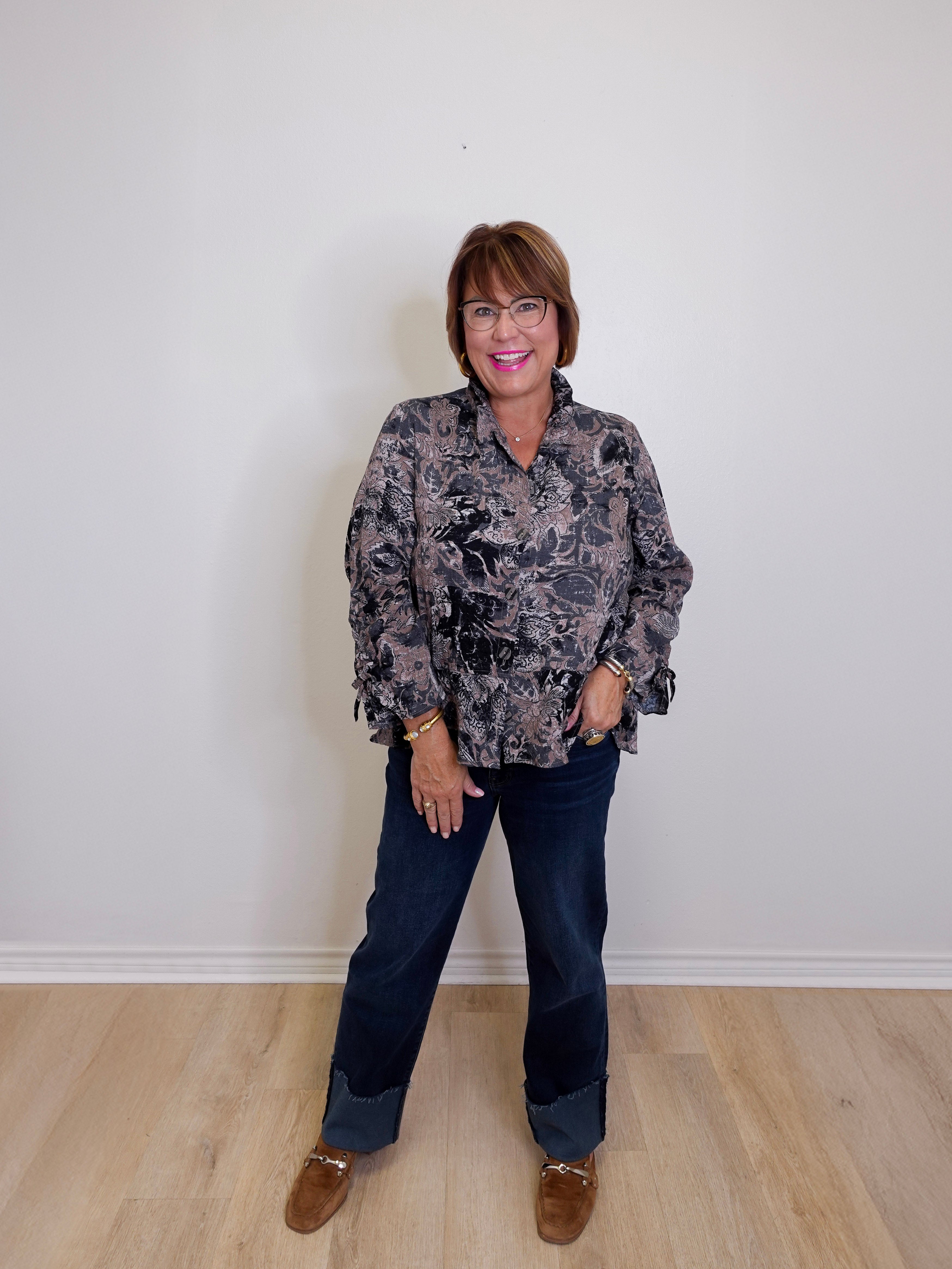Woman wearing a patterned blouse and jeans standing against a plain white wall.