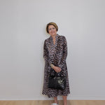 Woman wearing a leopard print dress with a black handbag against a plain white wall.