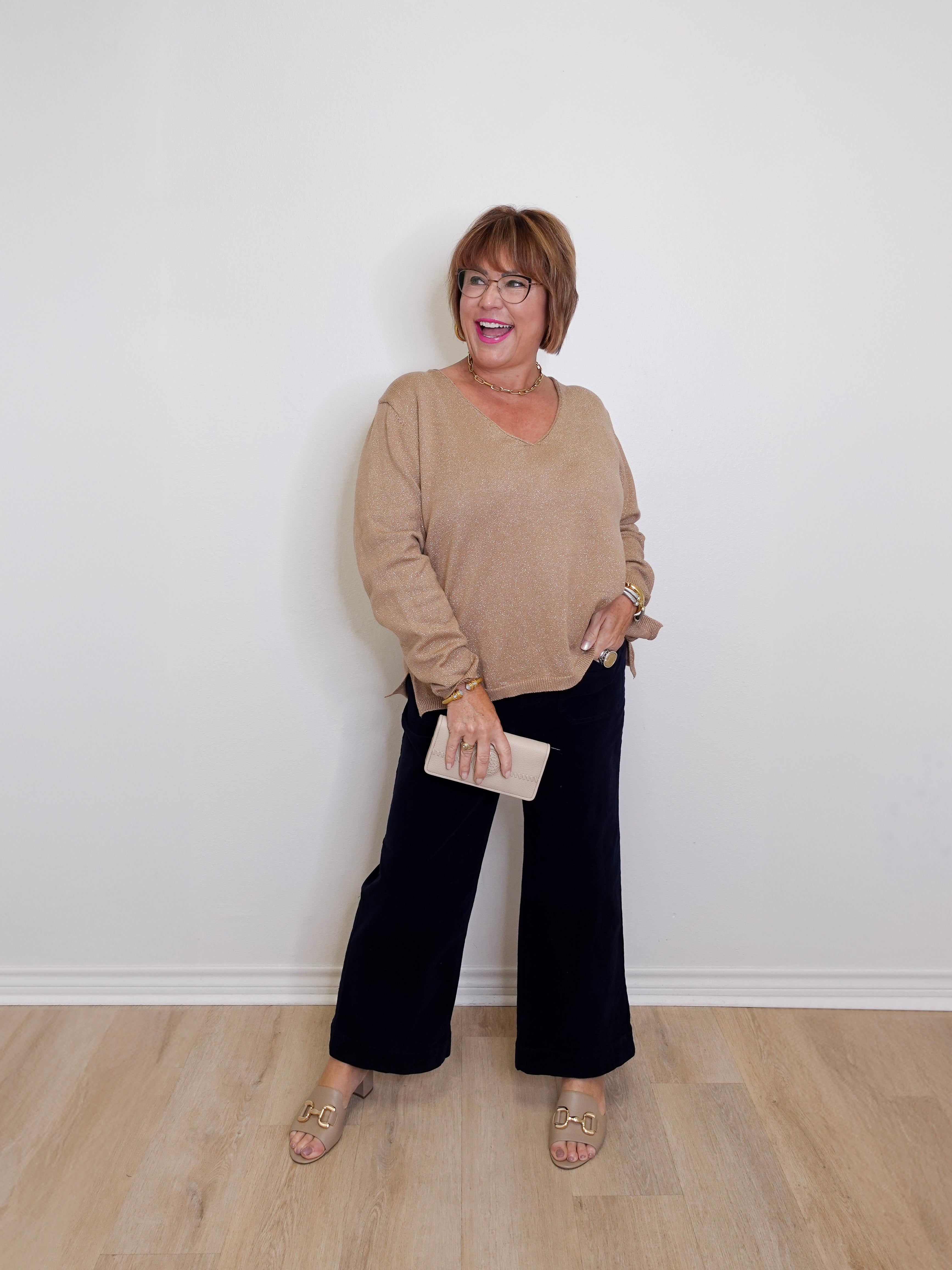 Woman wearing a beige sweater and black pants against a white wall.