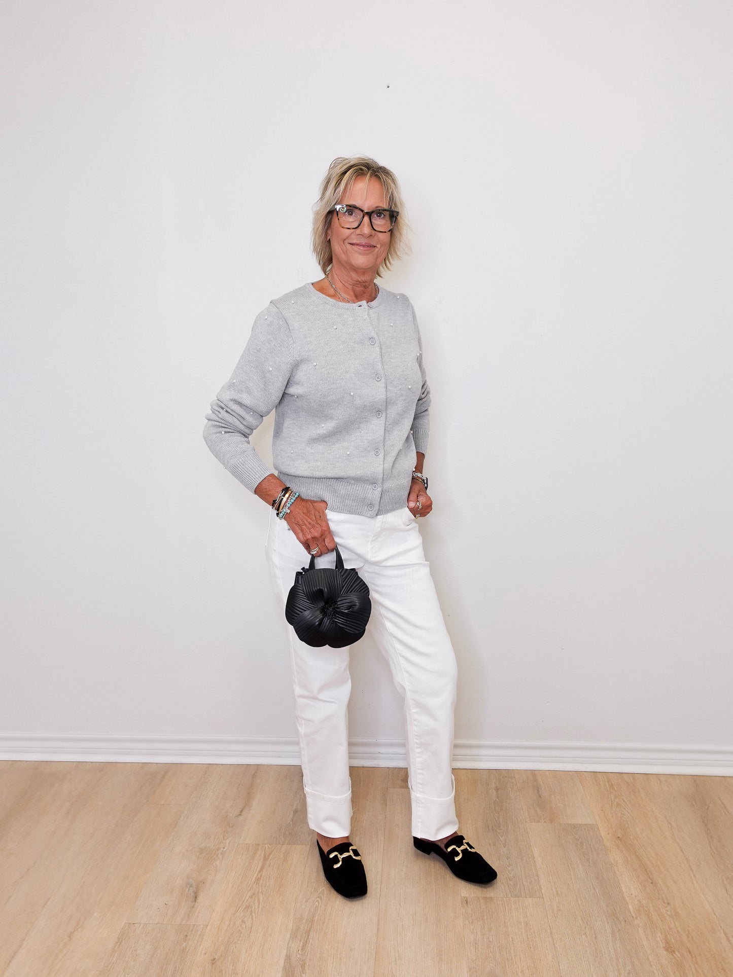 Woman wearing a gray sweater, white pants, and black shoes holding a black handbag against a white wall.