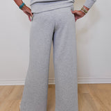 Person wearing a gray outfit with wide-leg pants against a white wall.