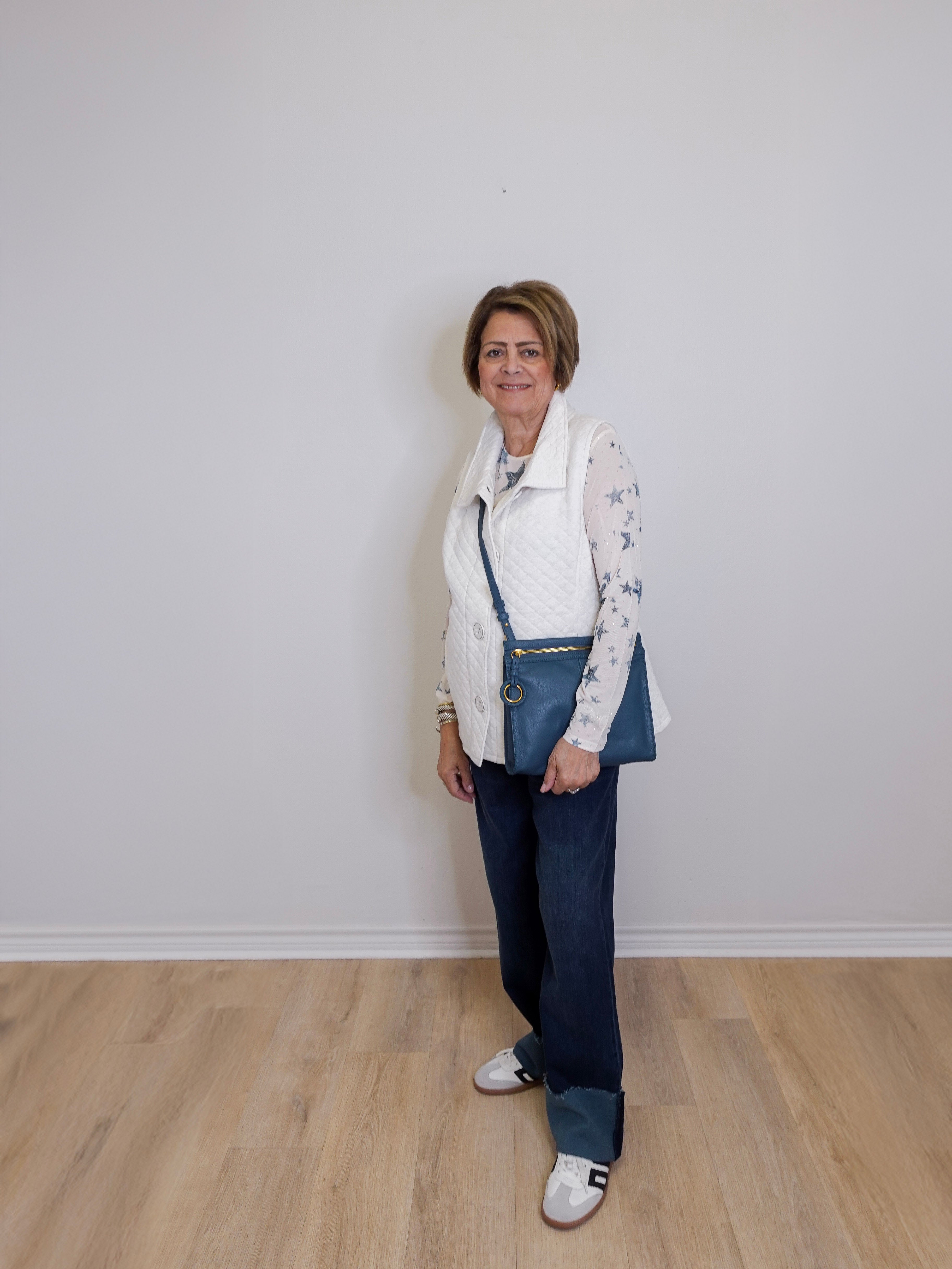 Woman wearing a white vest, patterned shirt, blue jeans, and white sneakers against a plain white wall.