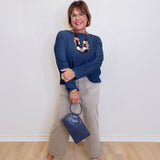 Woman wearing a blue sweater with a colorful necklace, beige pants, and holding a blue handbag against a white wall.