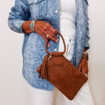 Person wearing a blue denim jacket and white pants, holding a brown handbag against a plain background