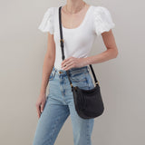 Person wearing a white top and blue jeans holding a black handbag against a plain background