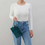 Person wearing a white long-sleeve top and blue jeans, holding a teal handbag against a gray background