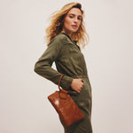 Woman holding a brown leather bag wearing an olive green outfit against a plain background