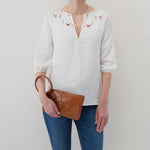 Person wearing a white blouse with floral embroidery and blue jeans, holding a brown leather clutch.