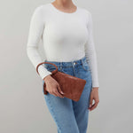 Person wearing a white long-sleeve top and blue jeans, holding a brown leather clutch against a plain background