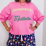 Person wearing a pink sweatshirt with 'Champagne & Mistletoe' text and patterned shorts.