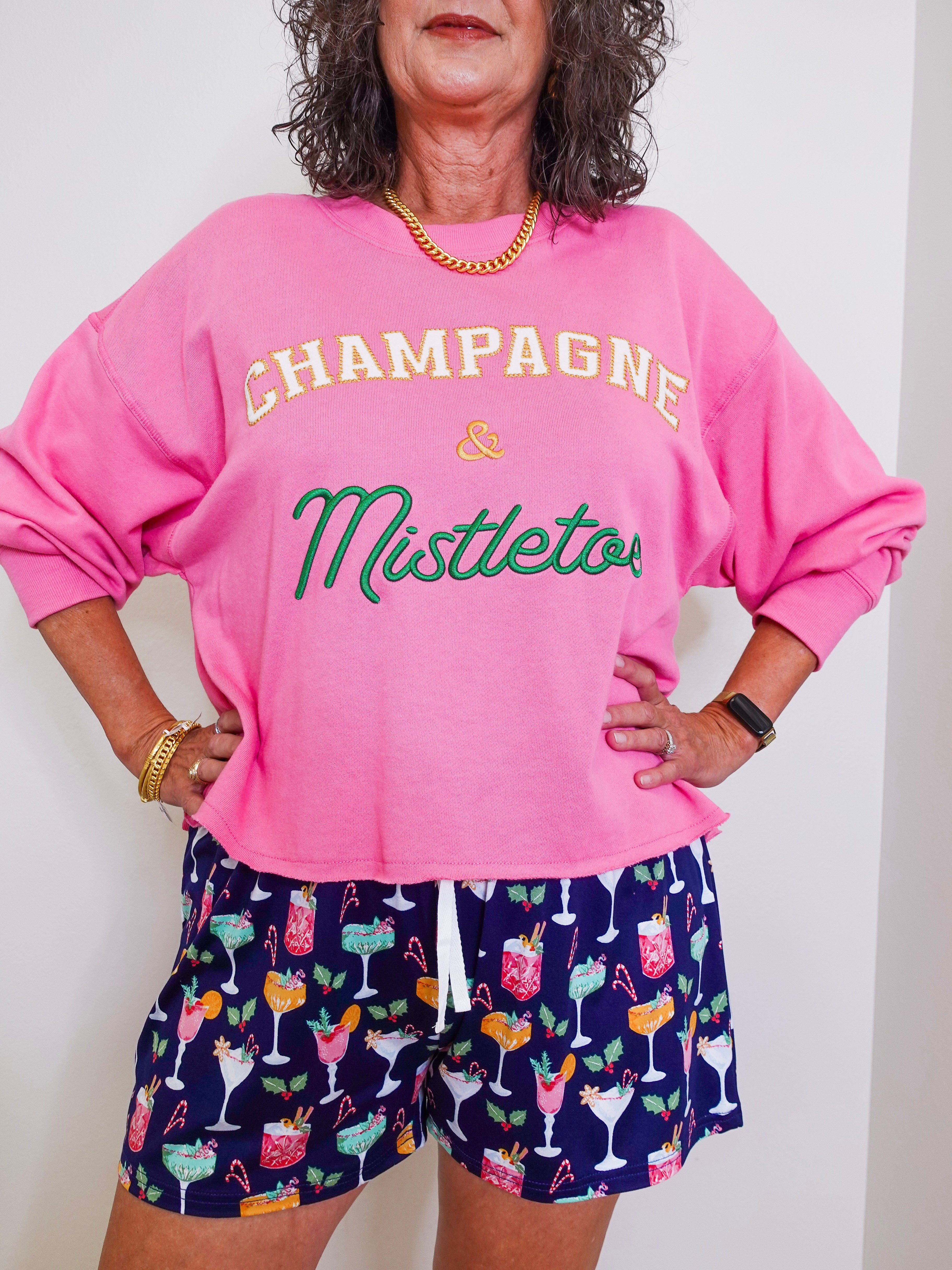 Person wearing a pink sweatshirt with 'Champagne & Mistletoe' text and patterned shorts.