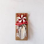 Metallic sugar scoop with peppermint candy on a cardboard holder against a white background