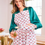 Woman in a kitchen wearing a patterned apron, holding a glass of wine and a bottle.