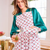 Woman in a kitchen wearing a patterned apron, holding a glass of wine and a bottle.