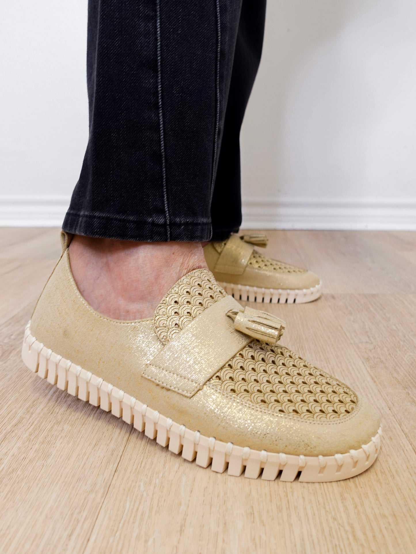 A person modeling a the ilse Jacobsen tassel loafer in gold against a white wall.