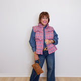 Woman wearing a pink plaid vest over a blue denim outfit, holding a brown handbag against a white wall.