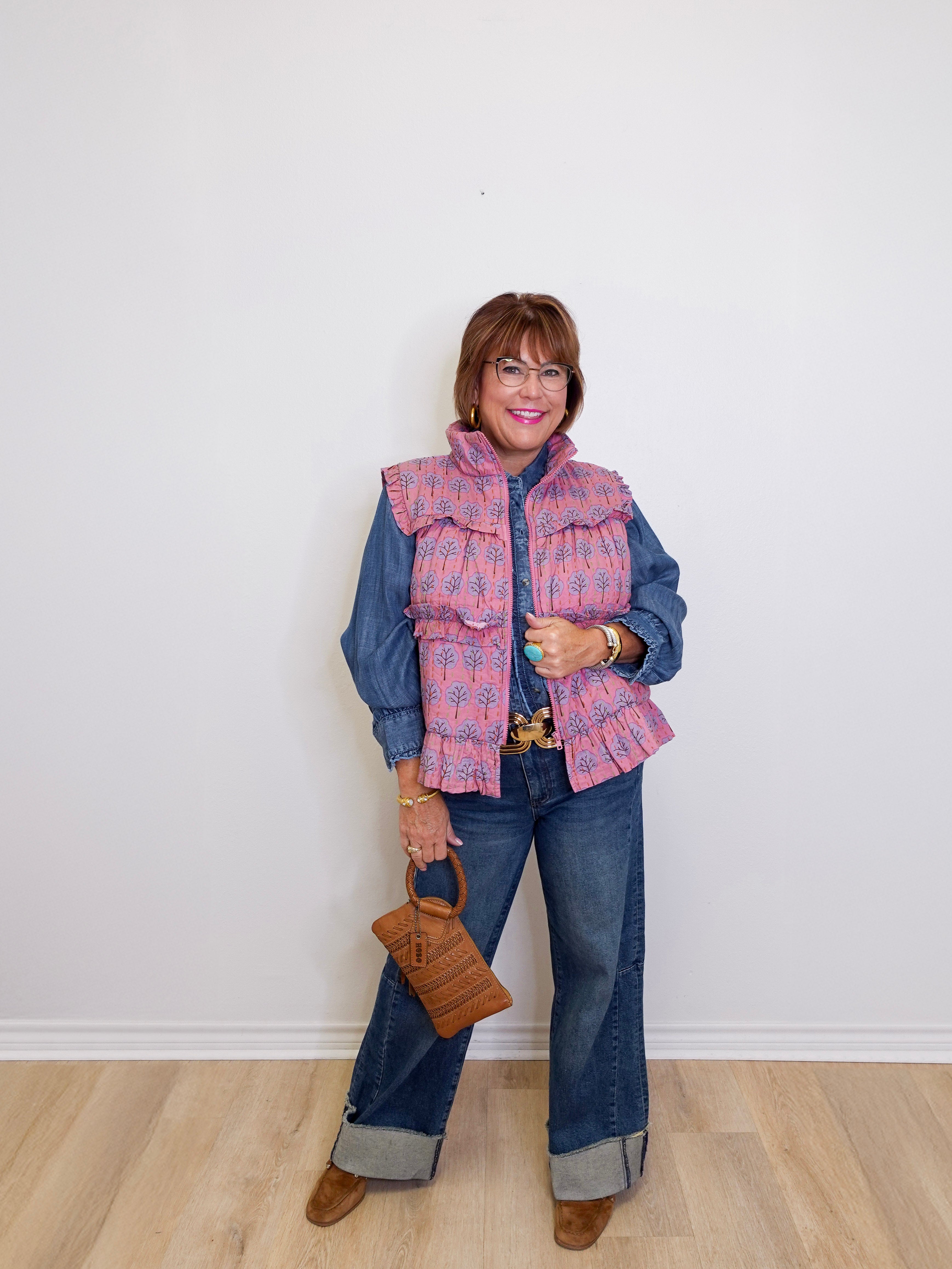 Woman wearing a pink plaid vest over a blue denim outfit, holding a brown handbag against a white wall.