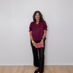Woman wearing a burgundy top and black pants holding a pink book against a white wall.