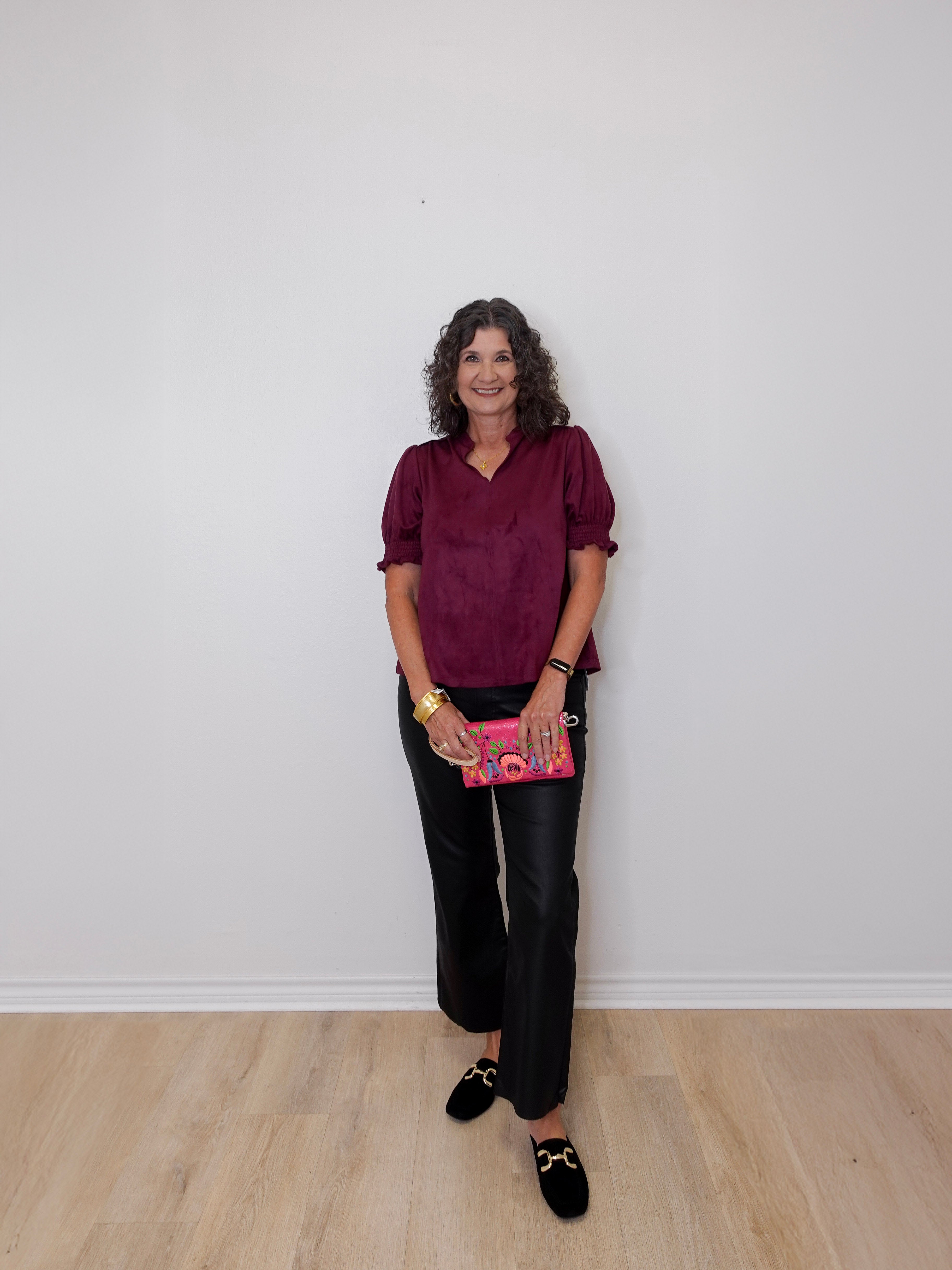 Woman wearing a burgundy top and black pants holding a pink book against a white wall.