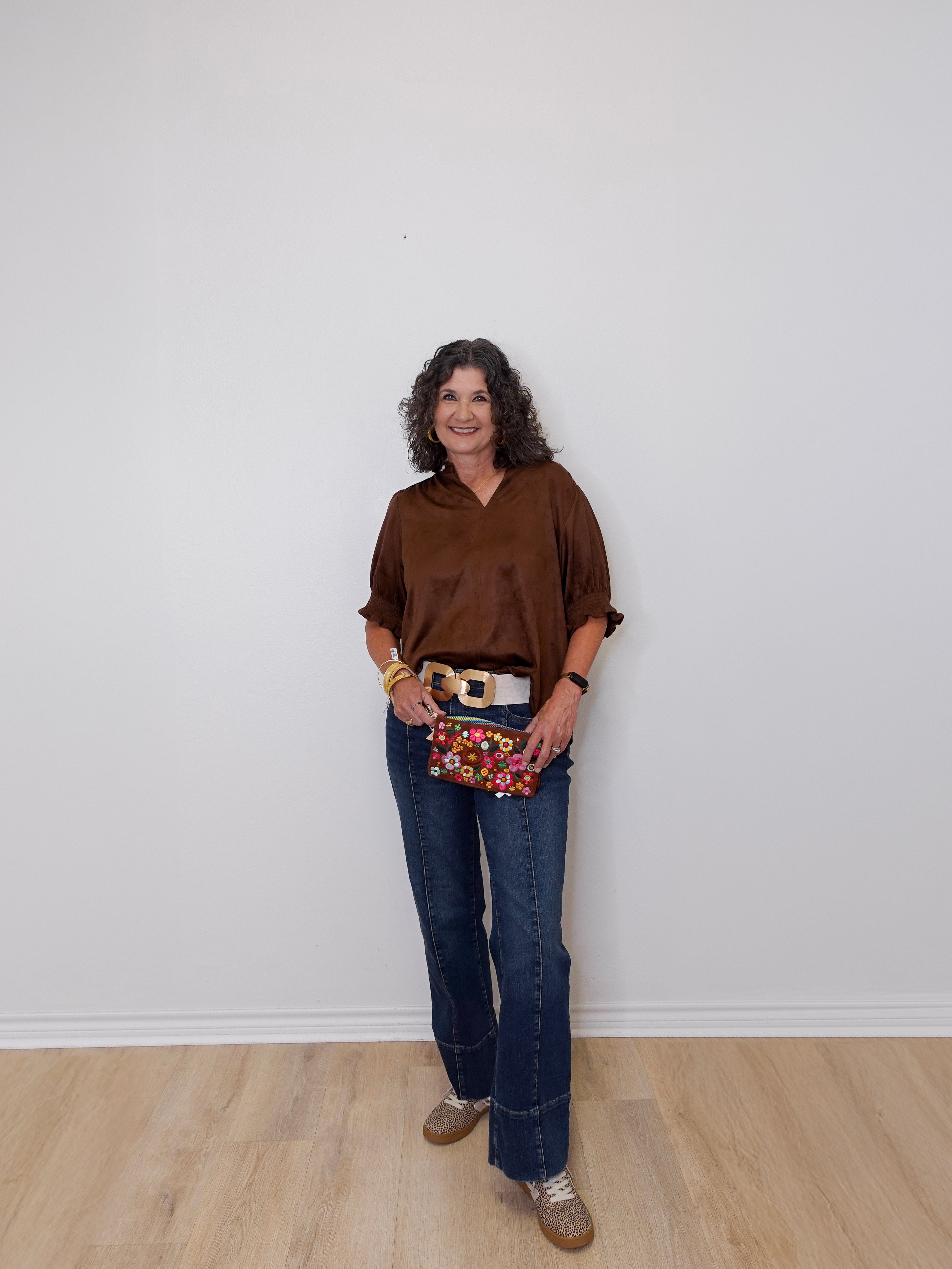 Woman wearing a brown shirt, blue jeans, and a patterned clutch against a white wall.