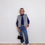 Person wearing a gray vest, navy sweater, blue jeans, and white sneakers against a white wall.