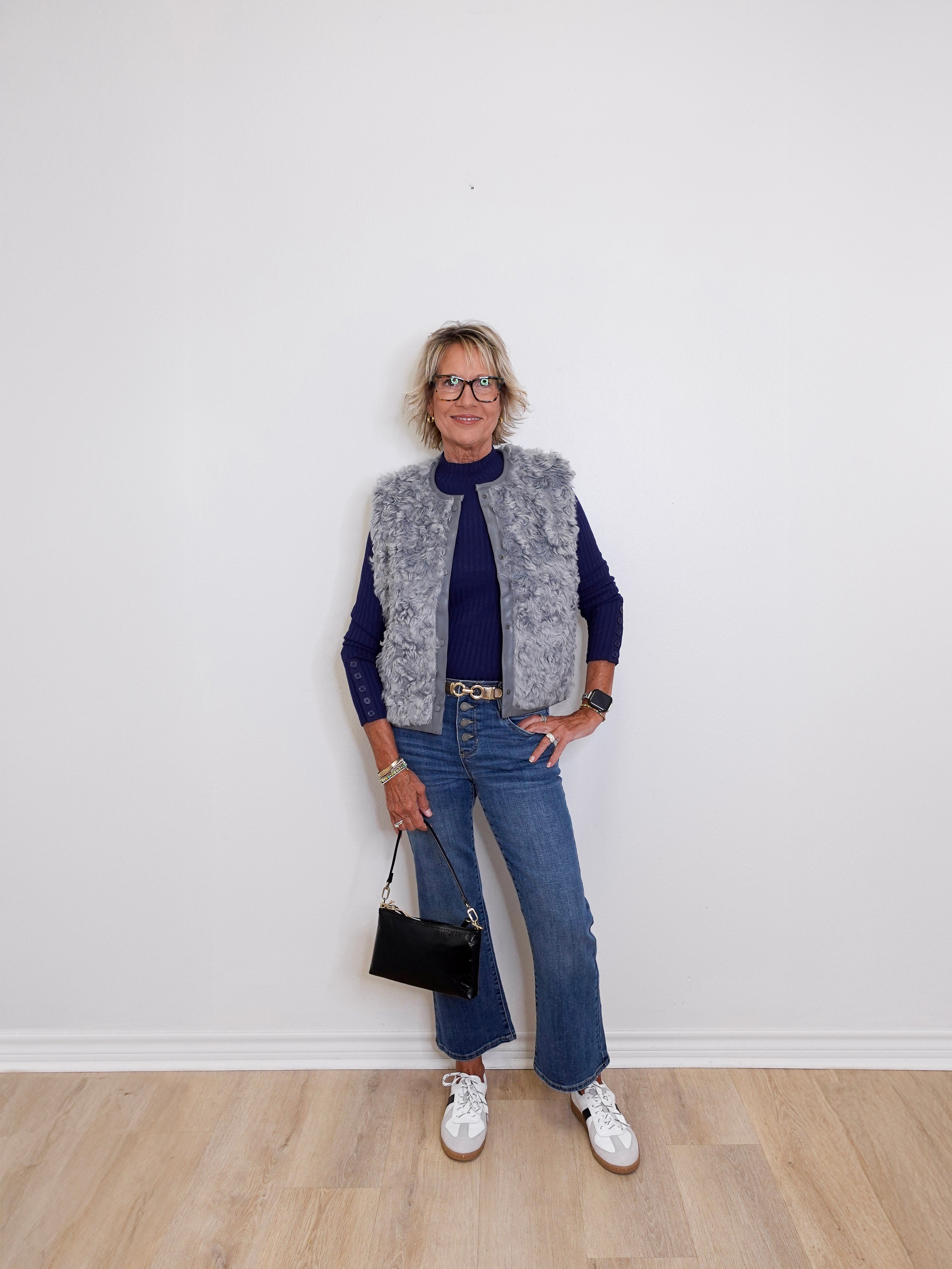 Person wearing a gray vest, navy sweater, blue jeans, and white sneakers against a white wall.