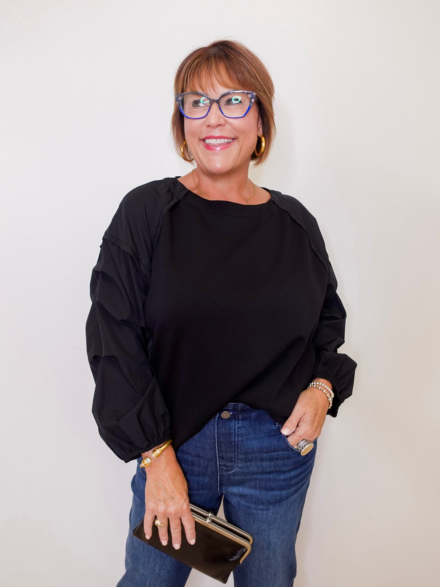 Woman wearing a black top and blue jeans, holding a clutch against a plain background