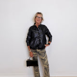 Person wearing a black leather jacket, camouflage pants, and white sneakers against a white wall.