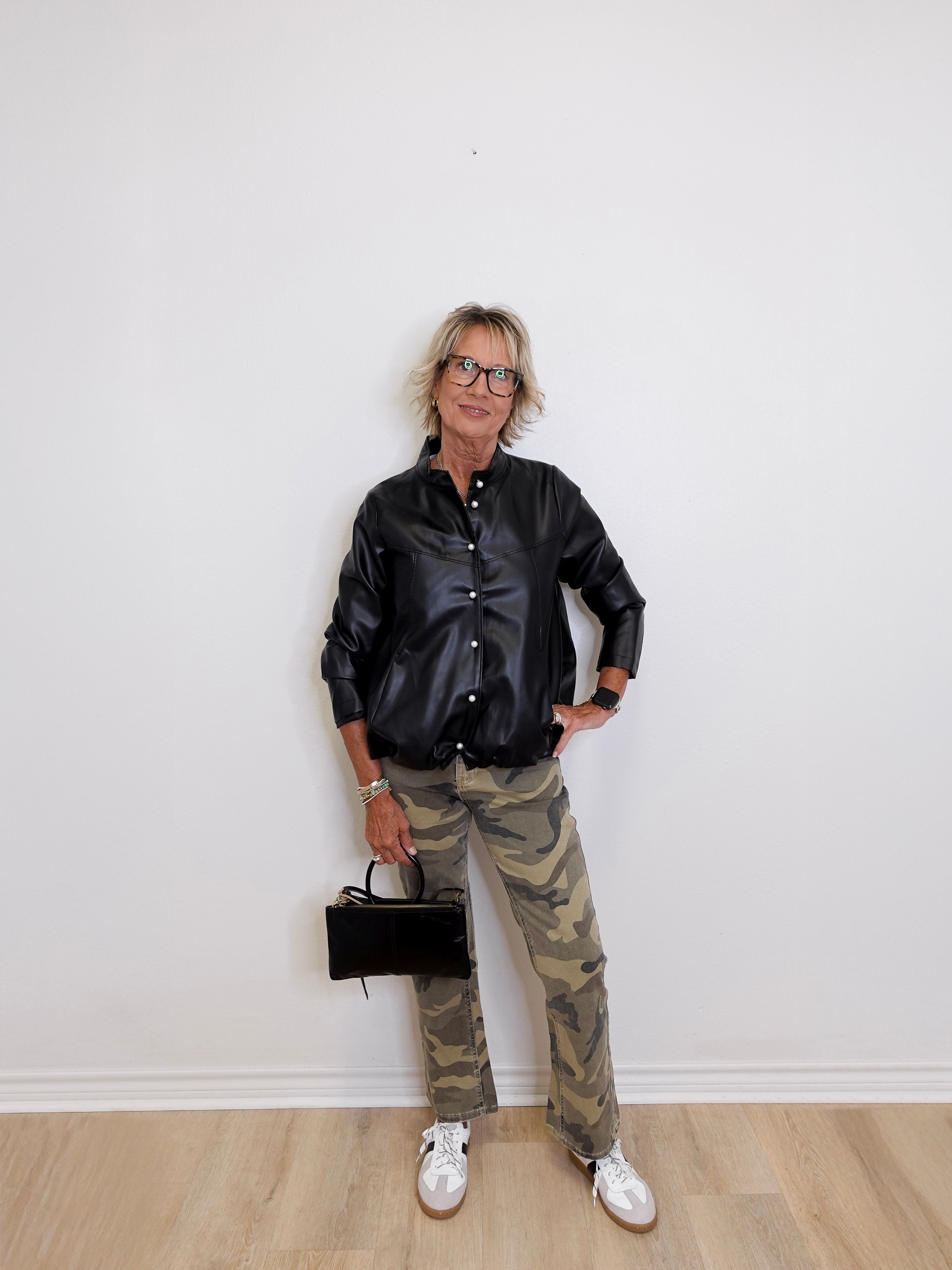 Person wearing a black leather jacket, camouflage pants, and white sneakers against a white wall.