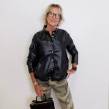 Person wearing a black leather jacket and camouflage pants holding a black handbag against a white wall.