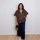 Woman wearing a brown vest and dark pants, holding a black bag with gold accents, standing against a white wall.