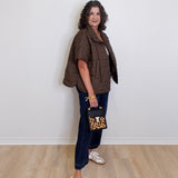 Woman wearing a brown jacket, blue jeans, and white sneakers holding a leopard print handbag against a white wall.