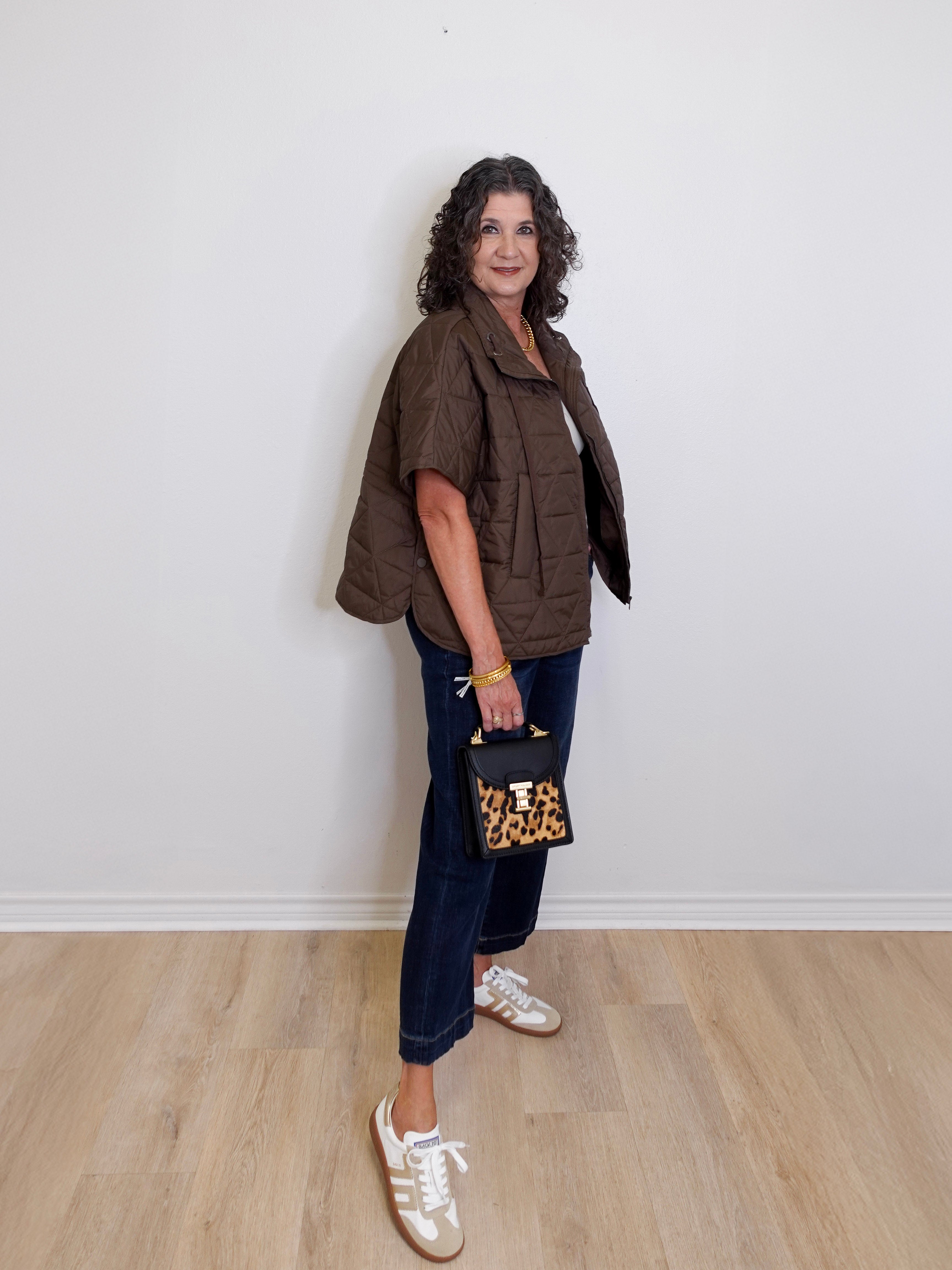 Woman wearing a brown jacket, blue jeans, and white sneakers holding a leopard print handbag against a white wall.
