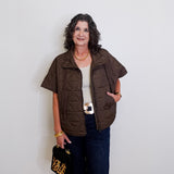Woman wearing a brown vest over a white top and dark pants, holding a leopard print handbag against a white wall.