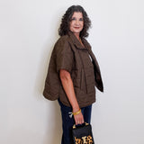 Woman wearing a brown puffer vest and holding a black handbag with leopard print accents against a white wall.