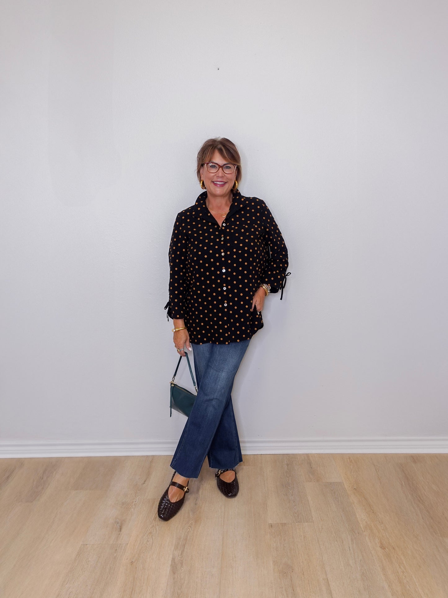Person wearing a black polka dot shirt and blue jeans against a white wall.