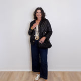 Woman wearing a black quilted jacket, white sneakers, and dark jeans against a plain white wall.